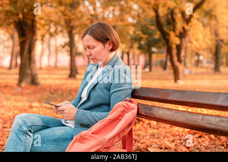 Woman typing text message on mobile phone sur banc de parc d'automne chaud d'automne, aux tons de couleurs image avec selective focus Banque D'Images