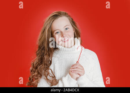 Joyeux Noël fille en pull blanc sur fond de couleur vive rouge vif. Portrait de la belle enfant. Copier l'espace. Cute blonde girl with sweet lollipop Banque D'Images