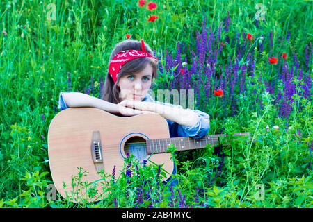 Belle fille avec une guitare dans les coquelicots. Jeune femme sur l'herbe. Un adolescent dans une veste en jean et un bandage rouge. Banque D'Images