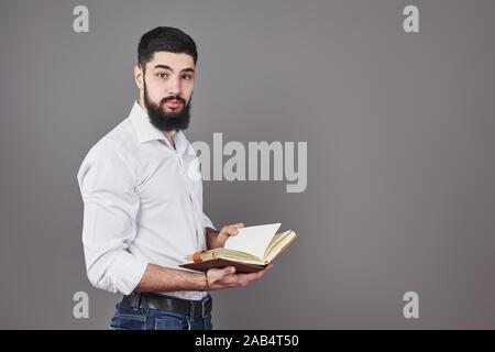 Portrait d'un jeune homme barbu portant une chemise blanche et la tenue d'un agenda ouvert et un stylo. Un mur gris Banque D'Images