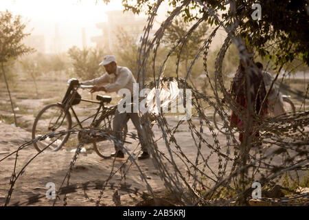 Un homme pousse son vélo de l'autre côté de bobines de fil de fer barbelé posé le long d'un chemin utilisé frquently à Kaboul, Afghanistan Banque D'Images