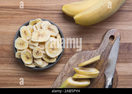 Frais et séchés, déshydratés chips de banane en noir bol sur planche de bois. Vue de dessus. Banque D'Images