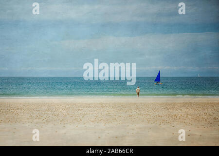 Image texturée d'un homme debout sur une plage donnant sur la mer, avec un bateau à voile bleu qui passe. Jersey, Channel Islands. Banque D'Images