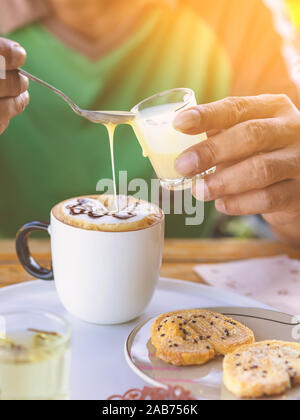 Un homme verser le lait dans une tasse de café à l'aide d'une cuillère pour éviter l'excès de douceur et mangé avec des biscuits couplé avec le thé chinois pour une collation en après-midi. Banque D'Images