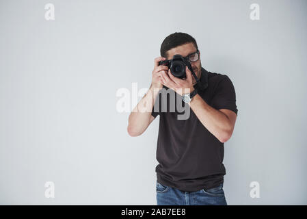 Un jeune homme hipster oculaires en détient un appareil photo DSLR en mains debout contre un mur blanc background Banque D'Images