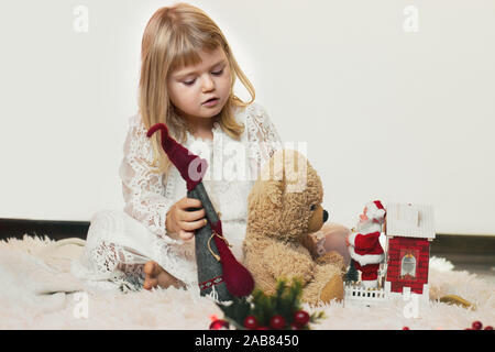 Jolie petite fille blonde assise sur le sol, jouant avec son ours en peluche et Petit Père Noël jouet de Noël Banque D'Images