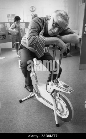 Années 1980, des cœurs sains, à l'intérieur d'un bureau, un employé exhuté, en pantalon et en cravate, s'est affaissé sur un vélo d'appartement d'intérieur, après avoir participé à l'initiative de son entreprise pour améliorer la santé de ses employés, Angleterre, Royaume-Uni. Banque D'Images