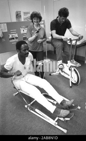 Années 1980, cœurs sains, à l'intérieur d'un bureau, une femme avec chronomètre supervisant deux employés masculins utilisant un kit de gymnastique à domicile, un rameur et un vélo d'exercice intérieur, dans le cadre d'une initiative pour améliorer la santé des gens, Angleterre, Royaume-Uni. Les années 80 ont été une décennie où l'accent a vraiment commencé à être mis sur la santé et le bien-être. Banque D'Images