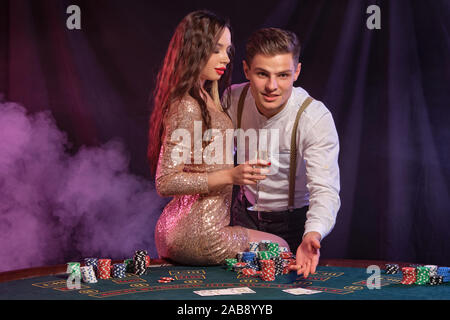 L'homme et la femme jouant au poker au casino, célèbre victoire à table avec des piles de jetons, cartes, champagne. La fumée noire, arrière-plan. Close-up. Banque D'Images