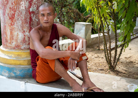 Un jeune moine bouddhiste avec crâne rasé habillé en costume orange et rouge repose par un pllar de son monastère le long de la rivière Chindwin, Myanmar (Birmanie) Banque D'Images