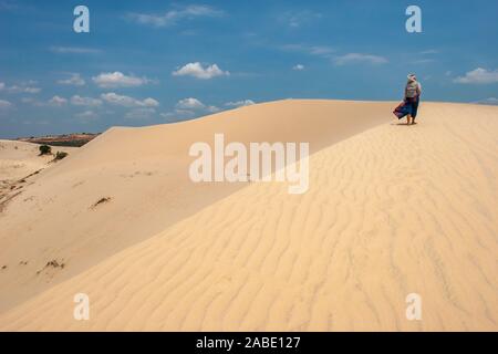 Une fille marche le long d'une grande dune de sable dans le vent. Jupe bleue s'agite. Capot, sur la tête. Banque D'Images