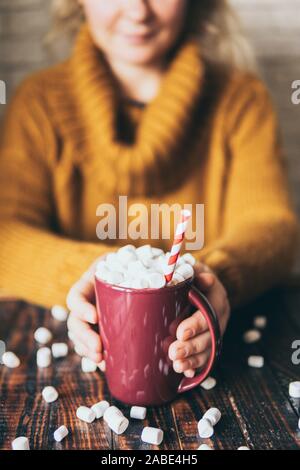 Femme en pull jaune holding mug de chocolat chaud recouvert de guimauve sur fond de bois sombre. Orientation verticale Banque D'Images