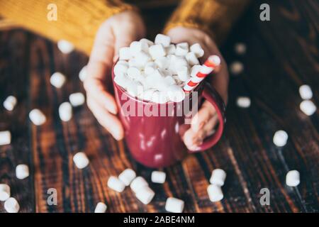 Femme en pull jaune holding mug de chocolat chaud recouvert de guimauve sur fond de bois sombre. Banque D'Images