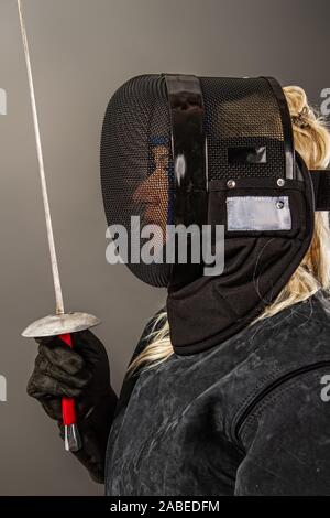 Jeune fille un tireur en noir et l'équipement d'entraînement d'un masque noir sur son visage se tenant une rapière pour préparer le combat sur un fond sombre. Banque D'Images