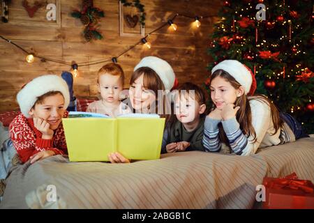 Mère lit un livre avec des enfants gisant sur le lit de la chambre avec un arbre de Noël. Banque D'Images