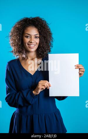 African American Woman holding white poster papier a4. Copier l'espace. Smiling pretty girl with curly hairstyle sur fond bleu. Banque D'Images