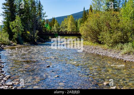 Belle montagne ruisseau coule sous un pont, avec un lit de rivière rocheux, des arbres et un ciel bleu. Banque D'Images