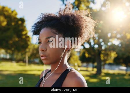 Close-up of a young african american woman standing in park à l'écoute de la musique via des écouteurs - déterminé jeune femme noire Banque D'Images