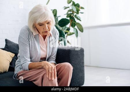 Senior woman having douleur au genou et assis sur un canapé dans l'appartement Banque D'Images