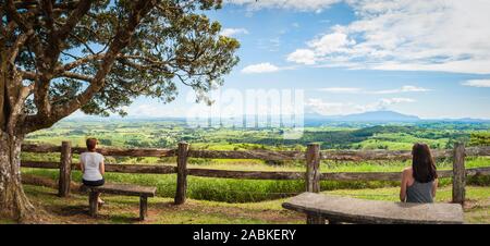 Paysage luxuriant des terres agricoles avec deux femelles assises sur un banc de pierre devant une clôture en bois rustique faite à la main au point de vue de Millaa Millaa, dans le Queensland. Banque D'Images