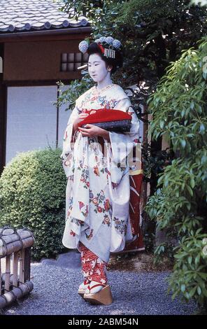 1987, historique, une jeune japonaise, maiko en vêtements ghesia traditionnels, avec son visage peint en blanc et les cheveux avec des fleurs décorées, elle porte une robe ornée, un kimono, avec un motif floral et des manches longues, des chaussettes boutonnées, avec des orteils fendus et de grandes sandales plates, connues sous le nom d'okobo. La maiko tient un sac kimono, un sac en tissu à fond plat avec cordon noué. Une maiko est une geshia plus jeune, une geshia apprentie encore en formation. Banque D'Images