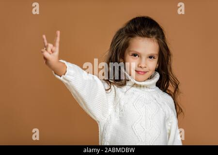Sourire mignon petit enfant fille en pull blanc faisant geste Rock sur fond beige. Banque D'Images