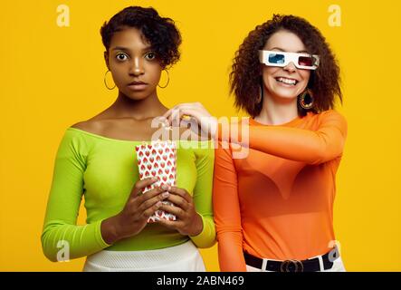 Portrait de deux jeunes filles dans le quartier branché d'hipster belle vêtements d'été colorés eating popcorn et regarder un film stéréo sur fond jaune. Banque D'Images