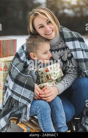 Mère et fils bénéficiant d'hiver neige journée en plein air, assis sur le traîneau, étreintes, couverts de plaid et holding Christmas gifts Banque D'Images