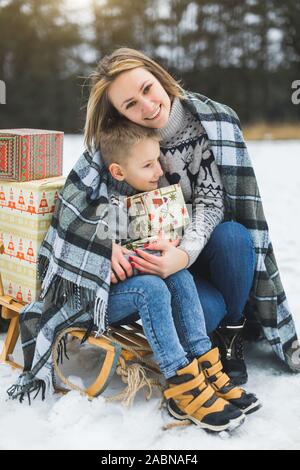 Heureuse mère et fils s'amusant avec traîneau dans une forêt d'hiver. Jour de neige d'hiver. Joli garçon avec sa maman sur un traîneau, serrant et souriante, tenant Banque D'Images