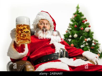 Père Noël bière potable près de l'arbre de Noël, les félicitant, l'air ivre et heureux. Modèle homme de race blanche en costume traditionnel. Nouvelle année 2020, des cadeaux, des vacances, de l'humeur d'hiver. Copyspace pour votre annonce. Banque D'Images