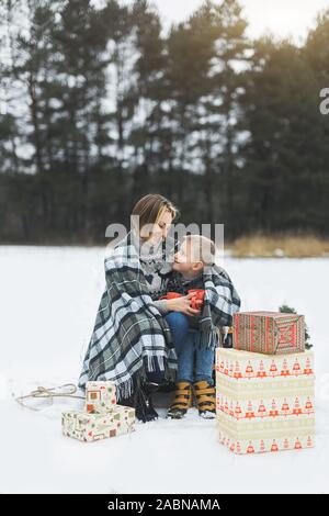 La mère et l'enfant assis sur le traîneau en bois en forêt d'hiver et de boire du thé chaud. Cadeaux de Noël présente sur la neige. La forêt enneigée sur l'hiver Banque D'Images