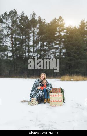 Mère et fils de passer du temps ensemble à la journée d'hiver. Mère et fils recouverts de plaid, tenant des tasses avec boissons chaudes, assis sur un traîneau décoré Banque D'Images