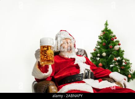 Père Noël bière potable près de l'arbre de Noël, les félicitant, l'air ivre et heureux. Modèle homme de race blanche en costume traditionnel. Nouvelle année 2020, des cadeaux, des vacances, de l'humeur d'hiver. Copyspace pour votre annonce. Banque D'Images