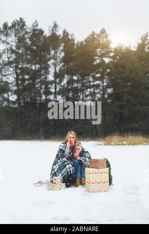 Mère et fils de passer du temps ensemble à la journée d'hiver. Mère et fils recouverts de plaid, tenant des tasses avec boissons chaudes, assis sur un traîneau décoré Banque D'Images