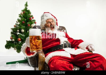 Père Noël bière potable près de l'arbre de Noël, les félicitant, l'air ivre et heureux. Modèle homme de race blanche en costume traditionnel. Nouvelle année 2020, des cadeaux, des vacances, de l'humeur d'hiver. Copyspace pour votre annonce. Banque D'Images