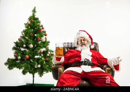 Père Noël bière potable près de l'arbre de Noël, les félicitant, l'air ivre et heureux. Modèle homme de race blanche en costume traditionnel. Nouvelle année 2020, des cadeaux, des vacances, de l'humeur d'hiver. Copyspace pour votre annonce. Banque D'Images