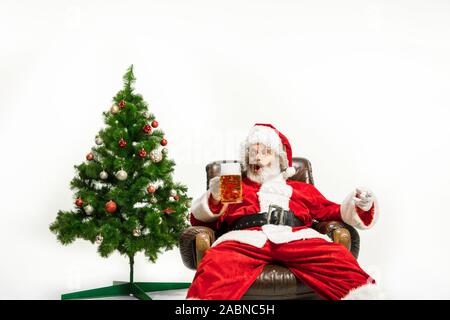Père Noël bière potable près de l'arbre de Noël, les félicitant, l'air ivre et heureux. Modèle homme de race blanche en costume traditionnel. Nouvelle année 2020, des cadeaux, des vacances, de l'humeur d'hiver. Copyspace pour votre annonce. Banque D'Images