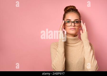 Une fille avec une peau propre et un haut bun se dresse sur fond rose et regarde dans le cadre, réglage de ses lunettes avec ses doigts. Banque D'Images