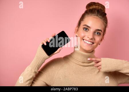 Une fille avec une peau propre et un haut bun joyeusement pointe vers le smartphone avec son doigt et regarde l'image. Banque D'Images