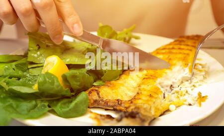 Les petits poissons de friture complet servi avec morceau de citron et feuilles de salade en plaque blanche. Femme est de manger du poisson avec fourchette et couteau en restaurant, close-up. Banque D'Images
