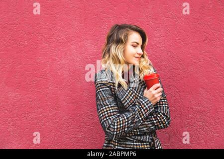 Portrait fille blonde avec les cheveux bouclés est titulaire d'une feuille de coin et une tasse de café pour aller dans ses mains. Sur fond rouge vif. Copier l'espace. Banque D'Images