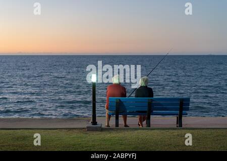 Les cheveux blancs âgés couple assis sur un banc à côté de la pêche en mer au coucher du soleil Banque D'Images