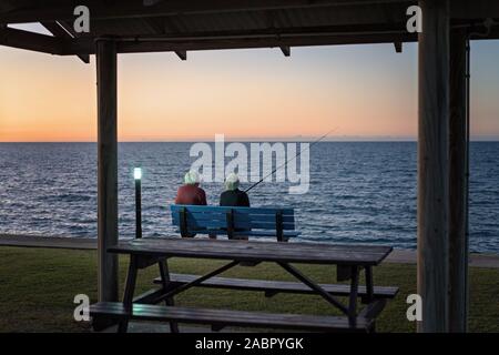 Les cheveux blancs âgés couple assis sur un banc en face d'une table de pique-nique à côté de la pêche en mer au coucher du soleil Banque D'Images