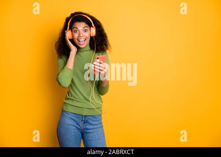 Photo de trendy ondulées bouclés chic fille positive joyeuse excitation exprimant millénaire sur le visage en denim jeans pull vert isolé dans le casque Banque D'Images