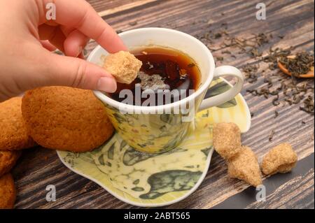 La main de la jeune fille met un morceau de sucre dans une tasse de thé noir, biscuits, les feuilles de thé, sucre brun sur un fond de bois. Close up Banque D'Images