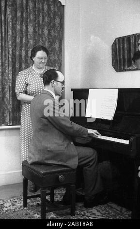 Années 1950, historique, à la maison une femme debout, regardant son mari jouer du piano, a Reval, Angleterre, Royaume-Uni. Avant la télévision, le divertissement à domicile était assuré par la radio, les magazines, les livres et les instruments de musique. Banque D'Images