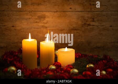 La chaleur de la notion de Noël : trois chandelles allumées sur une table en bois clair et un cadre rustique avec des branches de pins, décoration rouge et or et re Banque D'Images