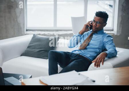 Photo de l'afro-américain jeune mec assis sur un canapé et avoir conversation de téléphone Banque D'Images