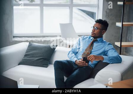 Regard de la réfléchie des lunettes. Guy Afroamerican assis sur le canapé et regardant son smartphone Banque D'Images