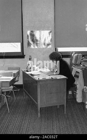 Années 1960, historiques, une femme travaillant dans un petit bureau à l'intérieur d'une chambre dans une université ou un collège étudiant en regard de certains bureaux en bois. Banque D'Images